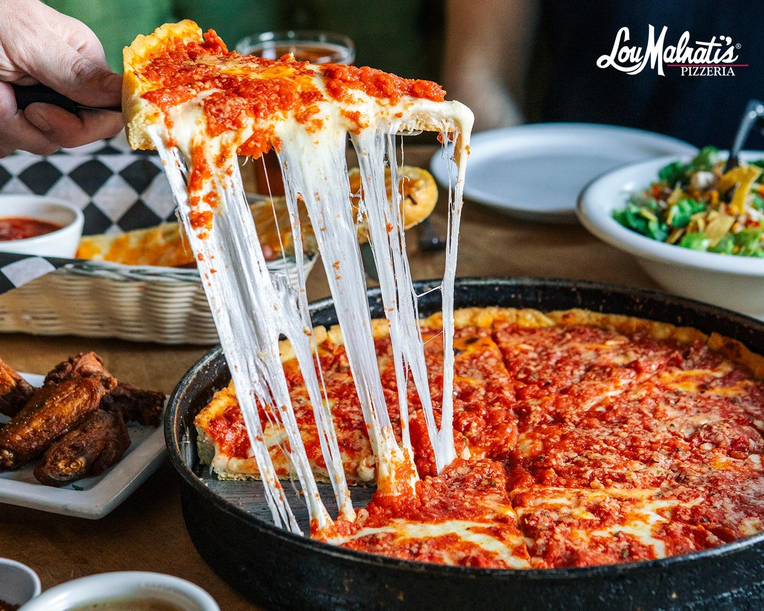 Lou Malnati's Pizzeria - A deep dish pan with a slice being pulled out by a hand. The cheese is stretching upwards from the pan in an impressive showcase of mozzarella. In the background are wings, a salad and cheesy garlic bread. . . 